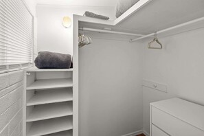 A white closet with shelves and hanging space, a small window with blinds, and a white wall.