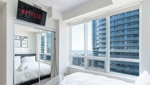 Une chambre d’hôtel moderne avec un grand lit, une armoire avec un miroir, un écran de télévision affichant « NETFLIX », et une grande fenêtre donnant sur la ville.