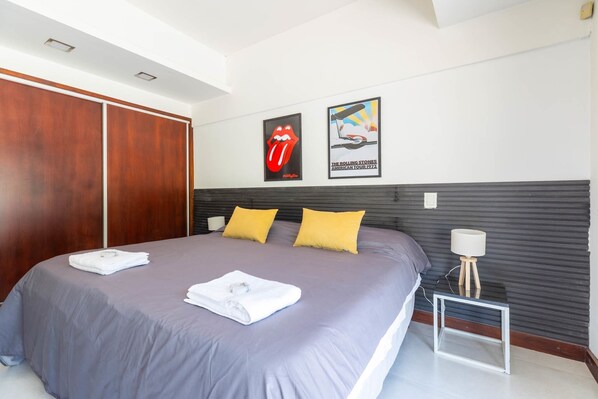 Una habitación de hotel con una cama grande, dos almohadas, una mesita de noche, una lámpara y dos carteles en la pared.