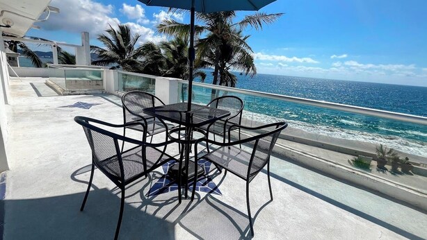 A sunlit outdoor seating area with a table and chairs, overlooking a beach with palm trees and the ocean. - OCEAN VIEW PENTHOUSE WITH TERRACE (Manzanillo)