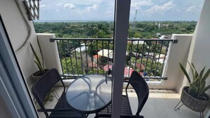 En balkon med et glasbord og to stole, med udsigt over et bybillede med grønt og bygninger.