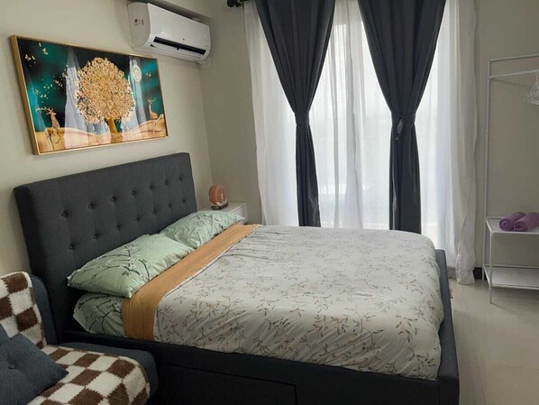 A bedroom with a bed, a nightstand, a wall-mounted air conditioner, a painting, and a window with curtains. - L L I Condotel (Lapu-Lapu)