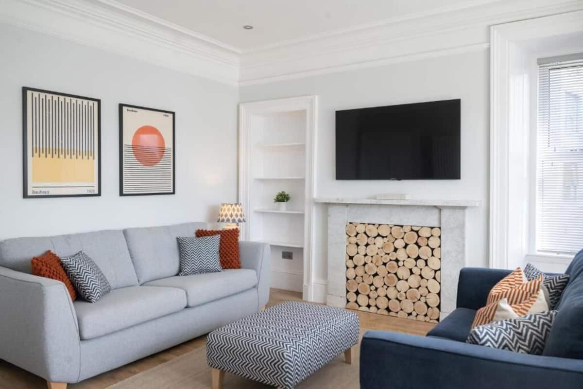 A modern living room with a grey sofa, blue armchair, and a patterned ottoman. A flat-screen TV is mounted on the wall, and a fireplace with a log pattern is visible.