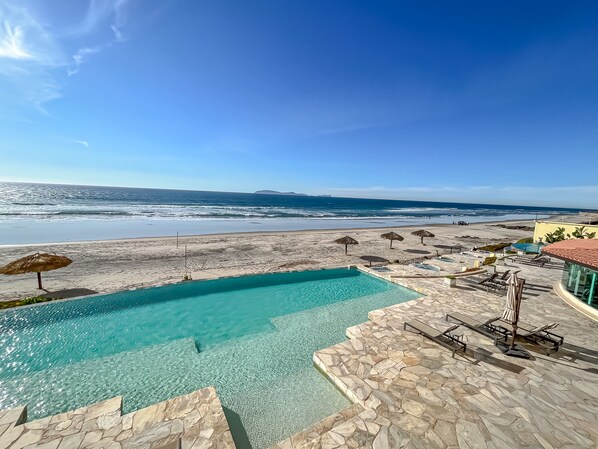 A clear blue sky, a swimming pool, and a beach with umbrellas.