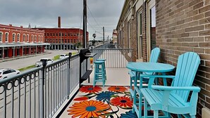 A balcony with a floral floor mat, two blue Adirondack chairs, and a view of a street with buildings. - Charming 2-bedroom condo located on the Historic Strand with WiFi, AC (Galveston)