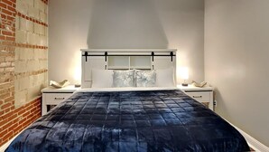 A bedroom with a brick wall, a bed with a dark quilt, two bedside tables with lamps, and a white headboard. - Charming 2-bedroom condo located on the Historic Strand with WiFi, AC (Galveston)