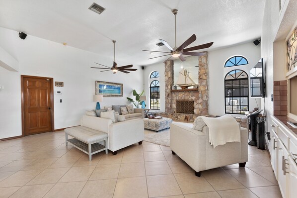 A spacious living room with a fireplace, two ceiling fans, and a view of the outside through large windows.