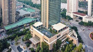 Un edifici alt amb façana moderna de vidre, envoltat d'altres edificis alts i una zona verda d'un parc.