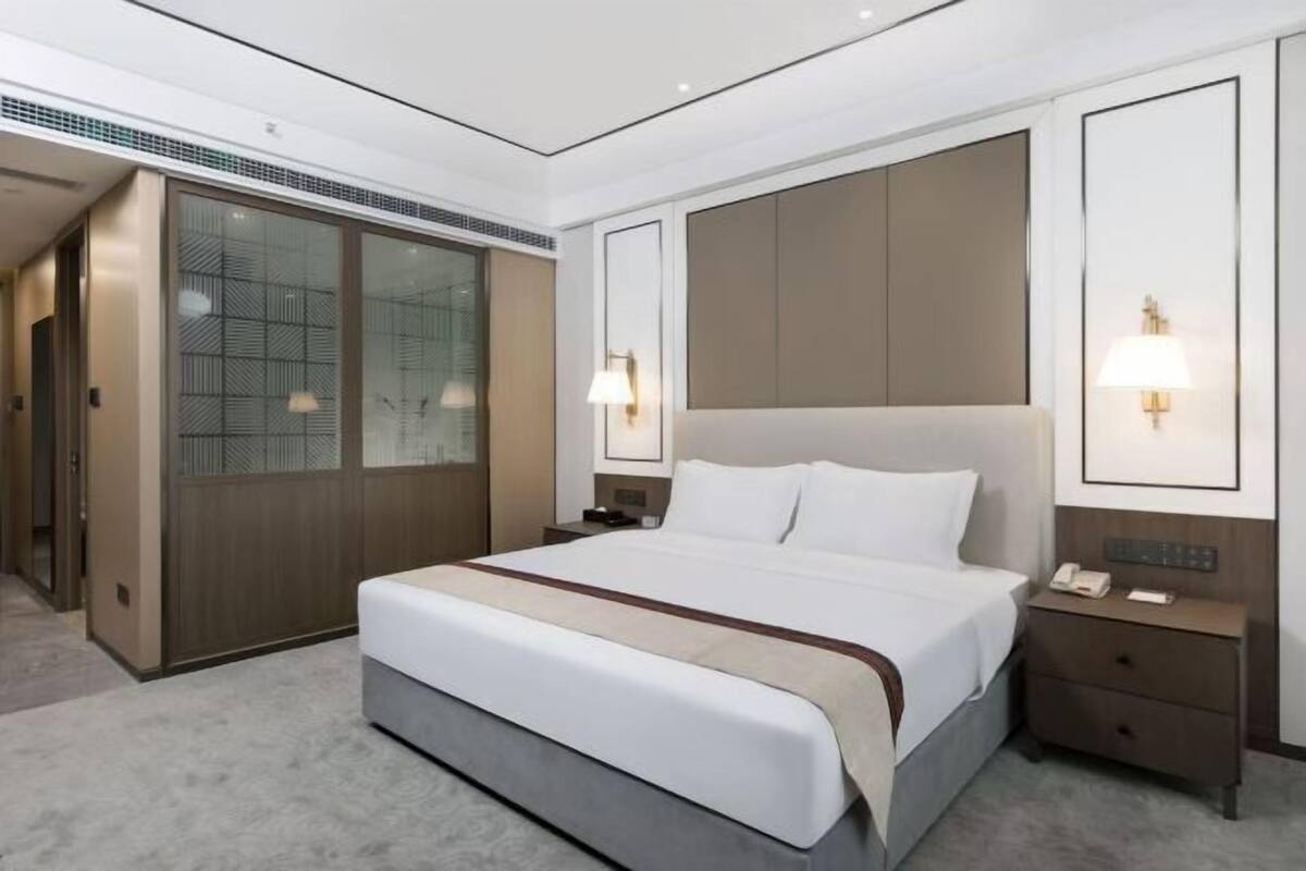 A modern hotel room with a large bed, a wardrobe, a bedside table with a lamp, and a wall-mounted phone.