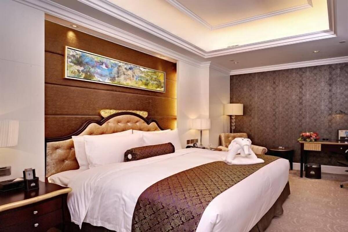 A hotel room with a large bed, a headboard with a painting, a desk, a chair, and a lamp.