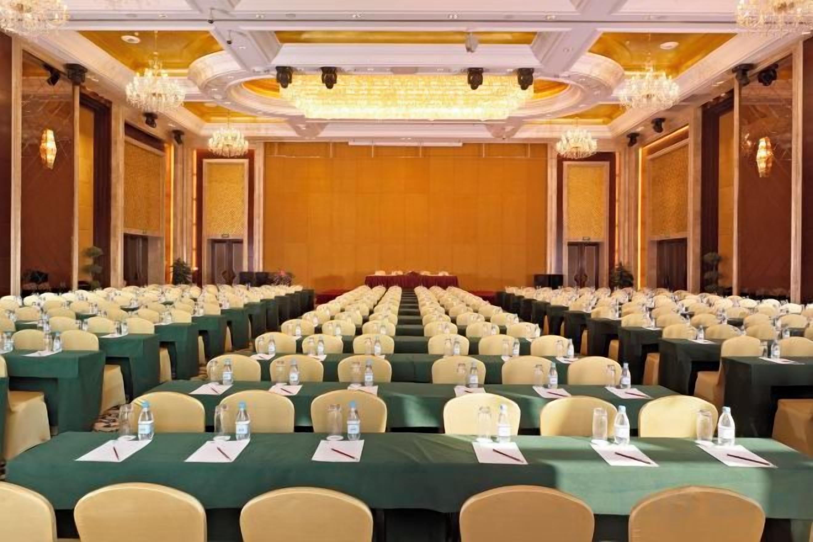 A conference room with rows of tables and chairs, a stage at the front, and chandeliers on the ceiling.