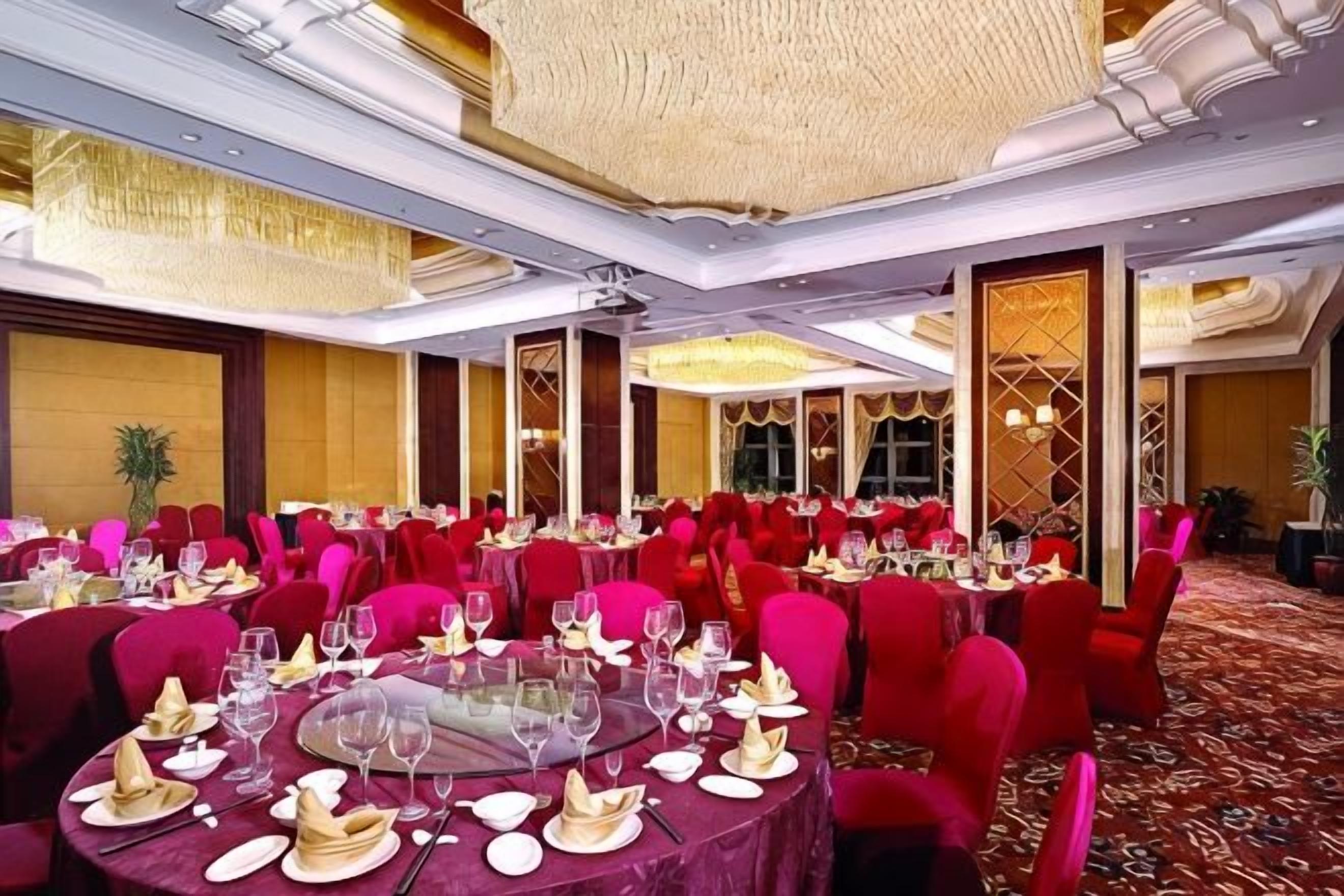Uma sala de banquetes com mesas redondas preparadas para refeições, cadeiras vermelhas e piso coberto por carpete.