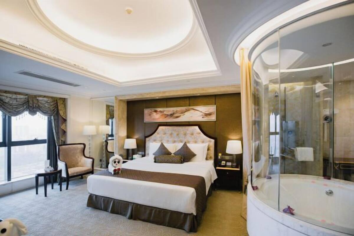 A hotel room with a large bed, a glass-enclosed shower, a desk with a chair, and a view of the city.