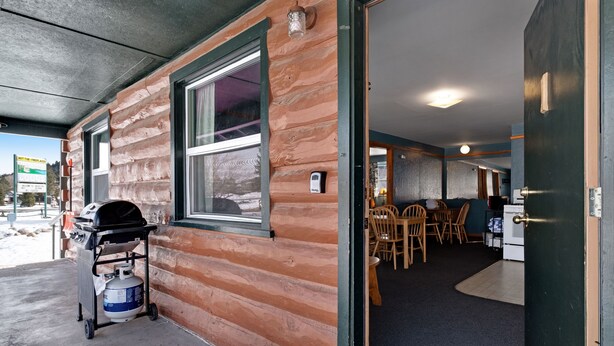 Il s’agit d’un bâtiment de style cabane en rondins, doté d’un patio comprenant un barbecue et une bouteille de propane. À l’intérieur, on trouve un coin repas avec des chaises et une table en bois.