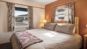 Een slaapkamer met een groot bed, twee ramen met bloemen gordijnen, een bedlampje en uitzicht op een besneeuwd landschap.