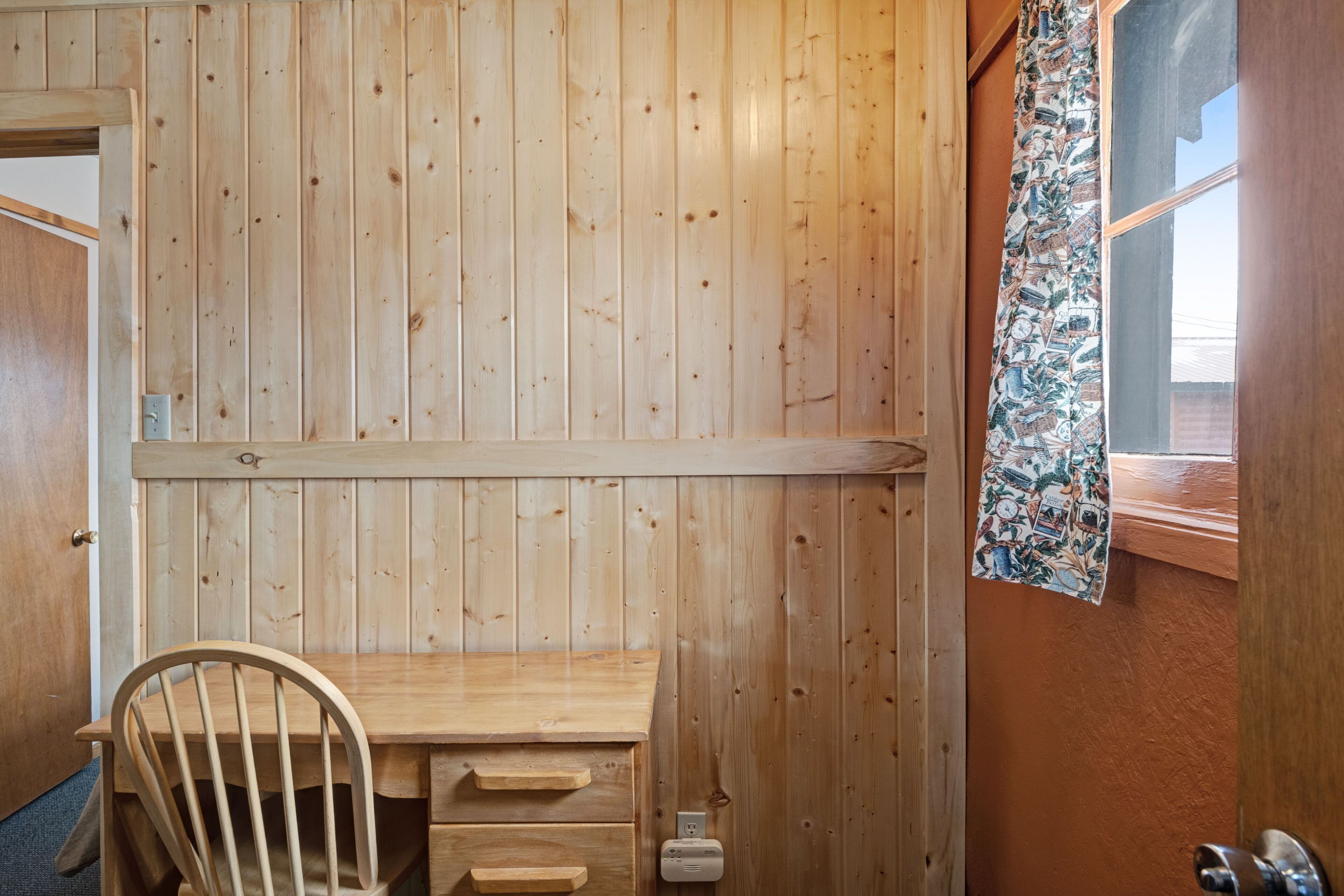 Una habitación de madera con un escritorio, una silla y un ventanal con cortinas estampadas.