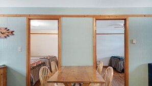 A room with a wooden dining table and chairs, a bed with a patterned blanket, and a small cabinet.