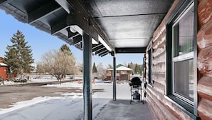 A covered porch with a concrete floor, wooden beams, and a window with a view of a snowy landscape and a building in the background.