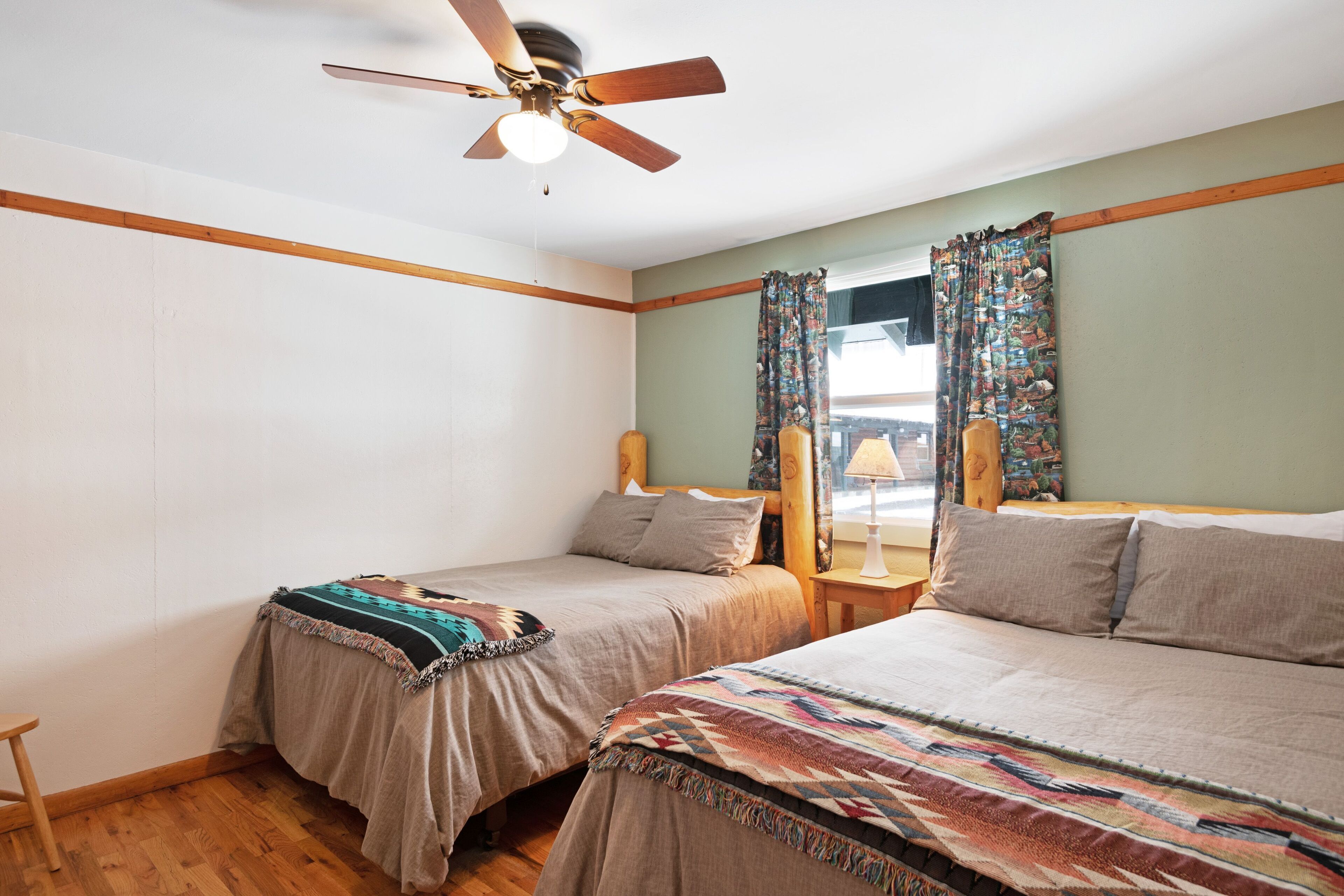 A bedroom with two single beds, a ceiling fan, wooden headboard, and a window with patterned curtains.