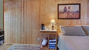 A wooden-paneled room with a bed, a nightstand with a lamp, a painting, and a radio. - Foothills Lodge 3 - South Fork (South Fork)