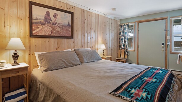 A wooden-paneled bedroom with a bed, two bedside lamps, a framed picture, and a door with a mirror. - Foothills Lodge 3 - South Fork (South Fork)