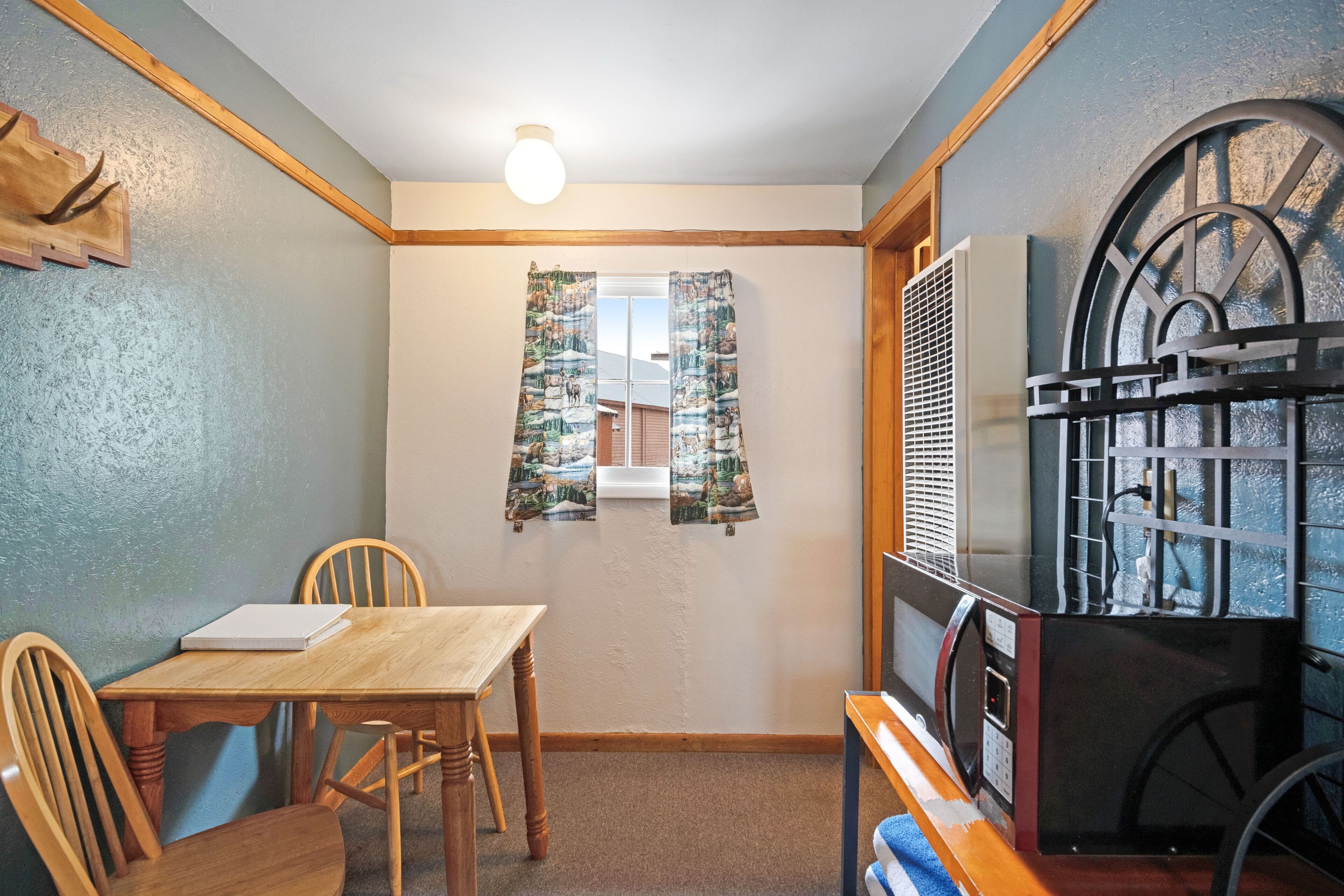 Una habitación pequeña con una mesa y sillas de madera, una ventana con cortinas, un radiador y un estante fijado a la pared.