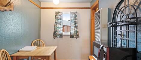 Una habitación pequeña con una mesa y sillas de madera, una ventana con cortinas, un radiador y un estante fijado a la pared.