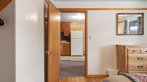 Una habitación con un aparador de madera, una cama con manta estampada y vistas a una cocina con armarios de madera y un refrigerador blanco.