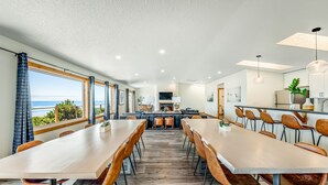 A modern dining area with a long table and chairs, a kitchen area with a dining table, and a view of the ocean. - Oceanfront, Hot Tub, Sleeps 23-62! (Waldport)
