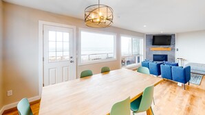 Een moderne woonkamer met een houten eettafel, groene stoelen, een blauwe zithoek en een grote televisie aan de muur.