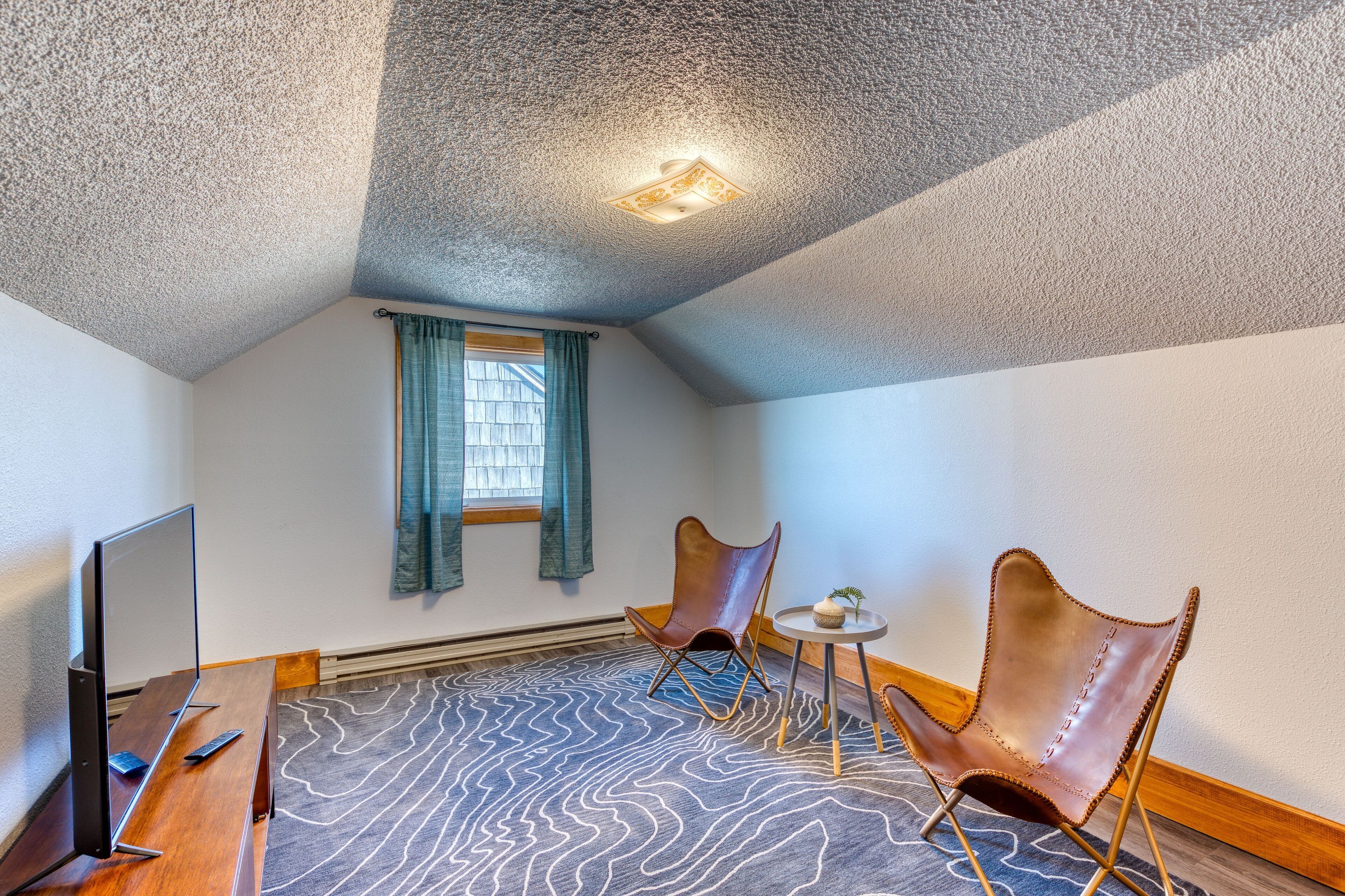 A small, cozy room with a sloped ceiling, a flat-screen TV, two leather chairs, a small table with a plant, and a patterned carpet.