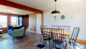Un comedor con una mesa y sillas de madera, un sofá azul y vistas al océano a través de amplias ventanas.