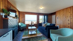 Una habitación acogedora con paredes de madera, un sofá azul, un sillón verde, una mesita y vistas al océano a través de amplios ventanales.