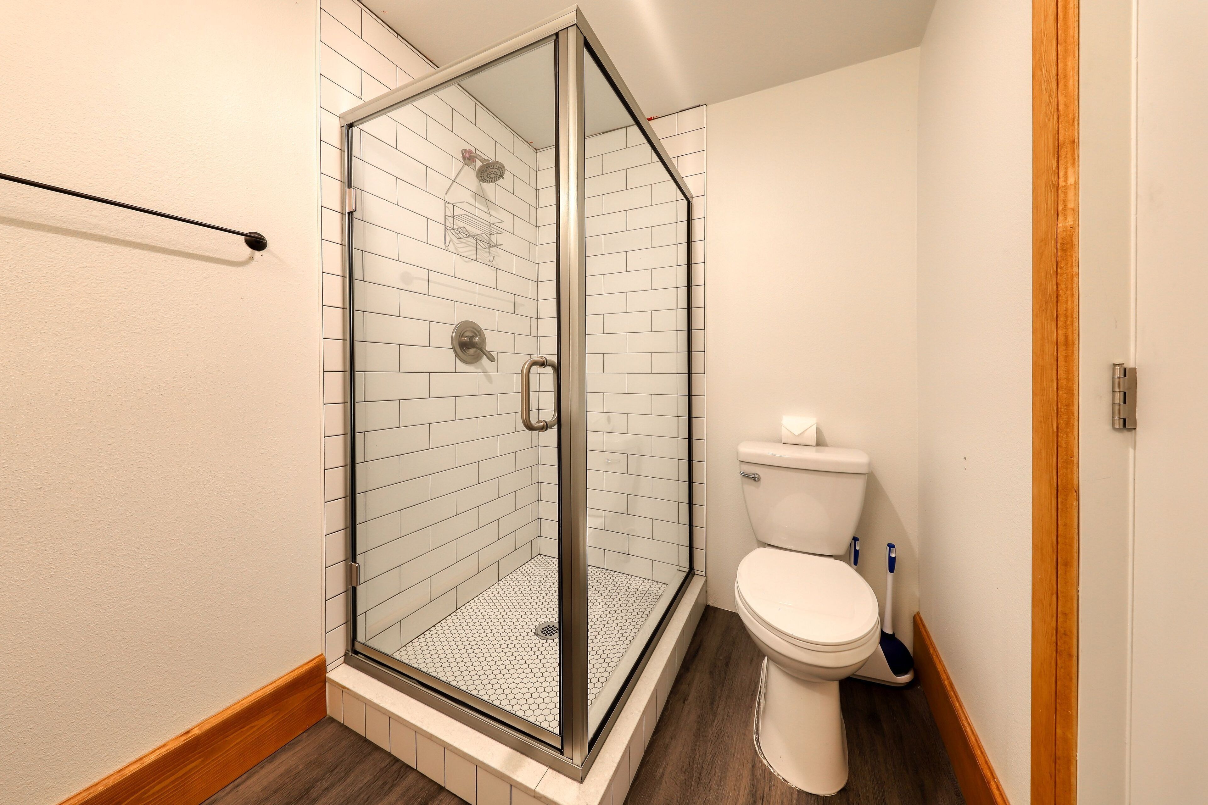 A bathroom with a glass-enclosed shower, white tiled walls, a white toilet, and a wooden floor.