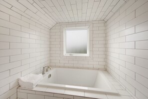 A modern bathroom with white tiled walls, a large bathtub, and a window above the sink.