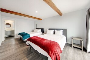 A modern hotel room with two beds, a wooden ceiling beam, a small bedside table, and a bathroom visible through an open door.