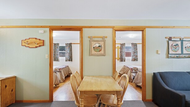 A dining area with a wooden table and chairs, a sofa, and a view through a doorway to a bedroom with a bed and curtains.