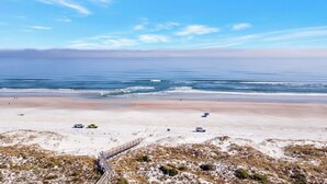 A beach with a wooden boardwalk leading to the water, a sandy area with sparse vegetation, and a clear blue sky.