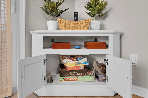 Un mueble blanco con una cesta tejida encima, que contiene juegos de mesa y un control remoto.