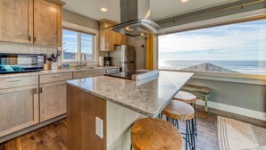 A modern kitchen with wooden cabinets, a large island with stools, a stainless steel refrigerator, and a view of the ocean. - Oceanside Inn #6: Pet-free unit. No pets allowed. (Oceanside)