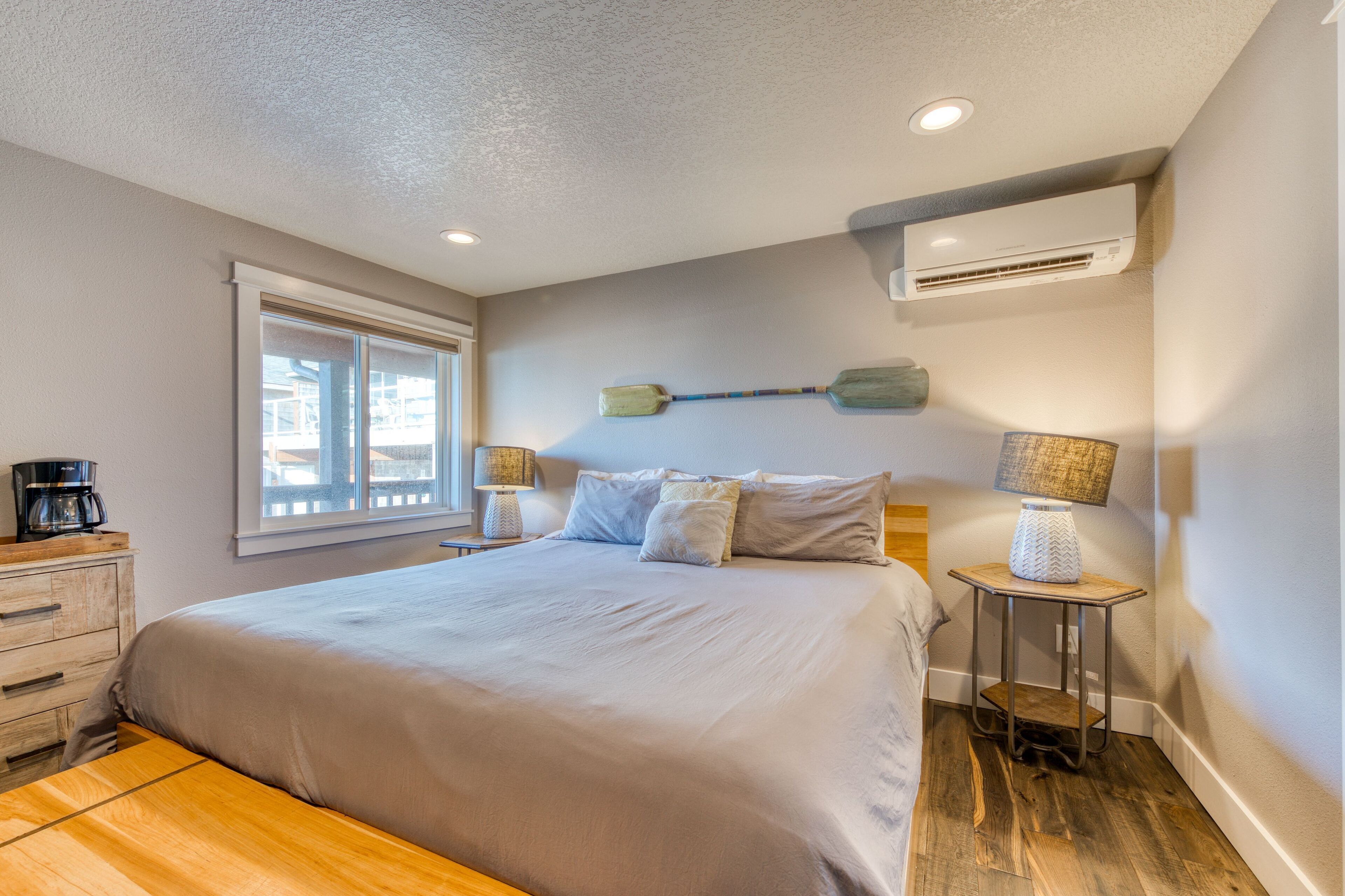 A bedroom with a large bed, a window, a nightstand with a lamp, and an air conditioning unit.