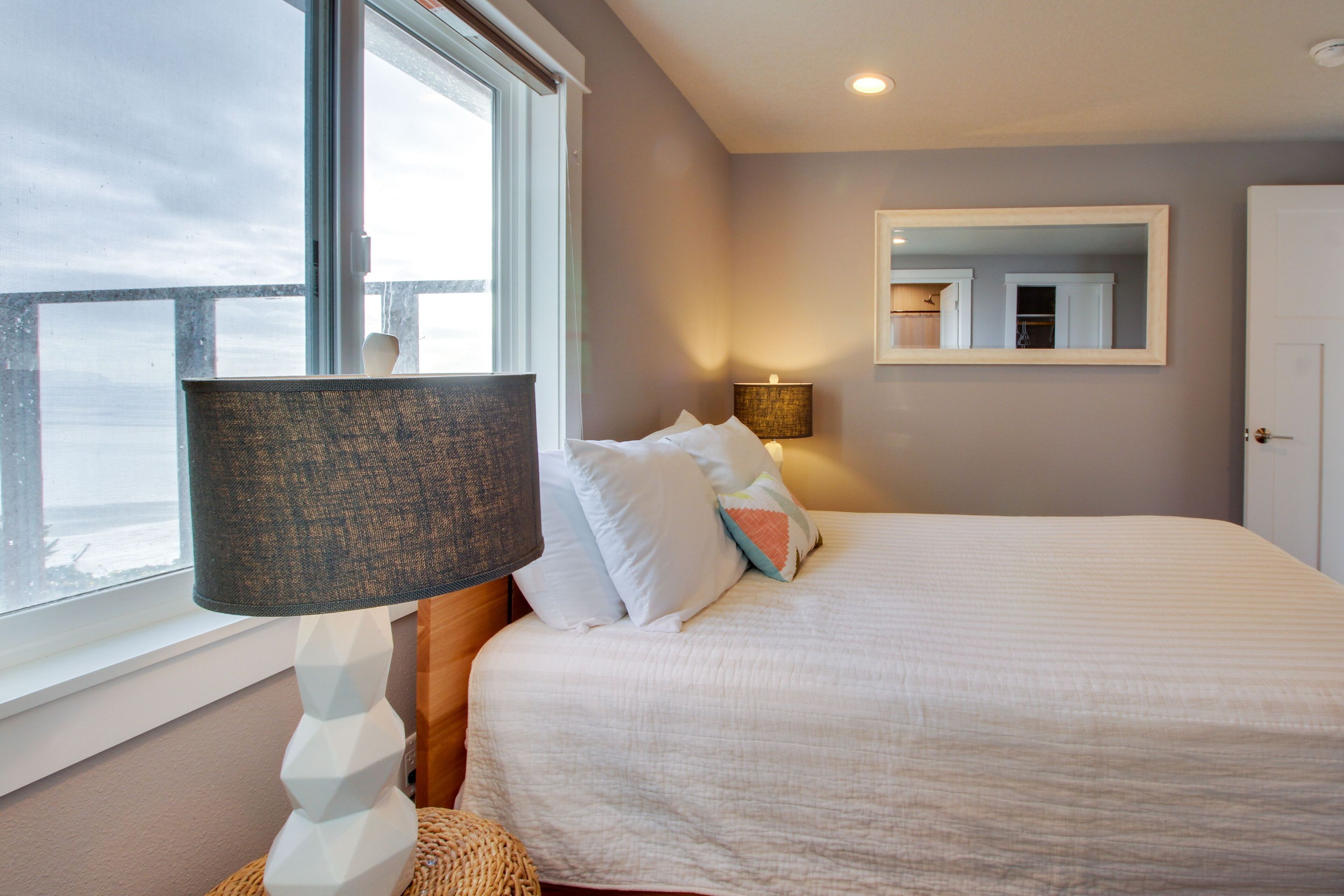 A bedroom with a bed, a lamp, a mirror, and a view of the sea.