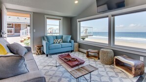 A living room with a sofa, a coffee table, and a TV. There are large windows with a view of the ocean.