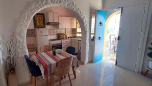 Zona pranzo con un tavolo apparecchiato per due, una cucina sullo sfondo e un'apertura che dà sull'esterno.