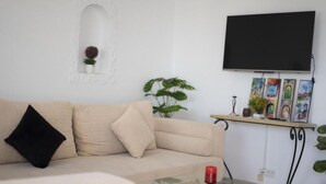 Un soggiorno con un divano color beige, un tavolino in vetro con una candela rossa, una TV montata a parete e una mensola con quadri incorniciati e piante.