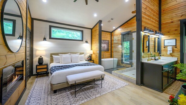 A modern bedroom with a large bed, bedside lamps, a fireplace, and a bathroom with a glass shower enclosure and wooden walls. - Couples ONLY + Spa Suite + Hot Tub + 3 Fireplaces - Escape to Whiskey Nights (Broken Bow)