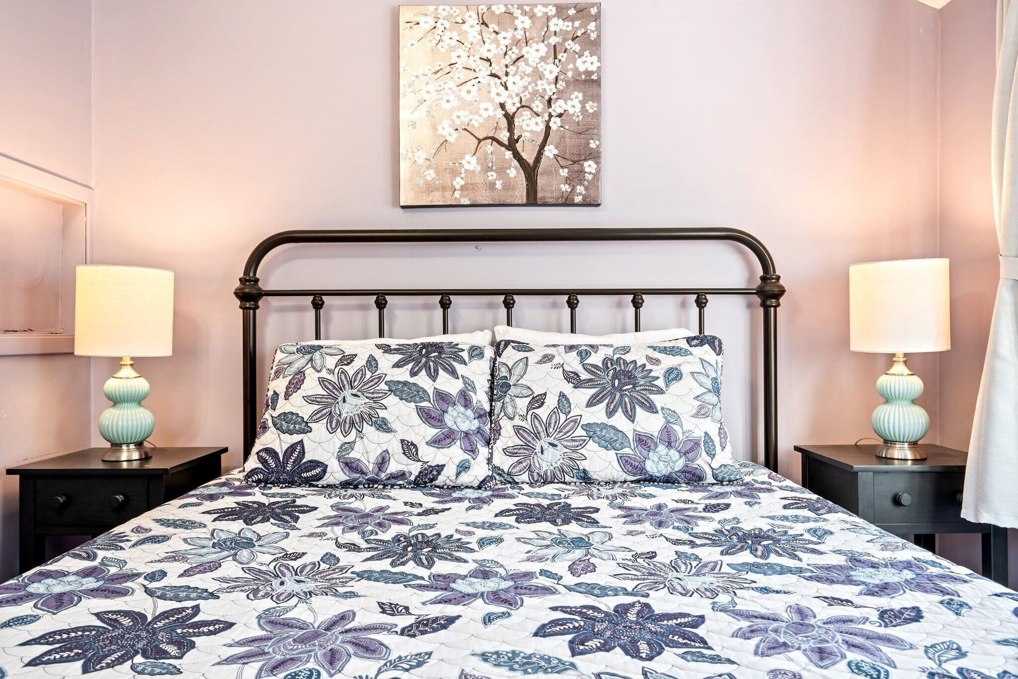 A bed with floral bedding, two bedside tables with lamps, a wall-mounted picture of a tree, and a mirror on the left wall.