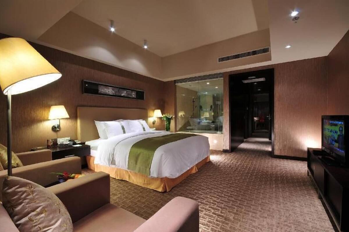 Habitación de hotel con cama, escritorio, silla, televisor y baño visible a través de una puerta de cristal.