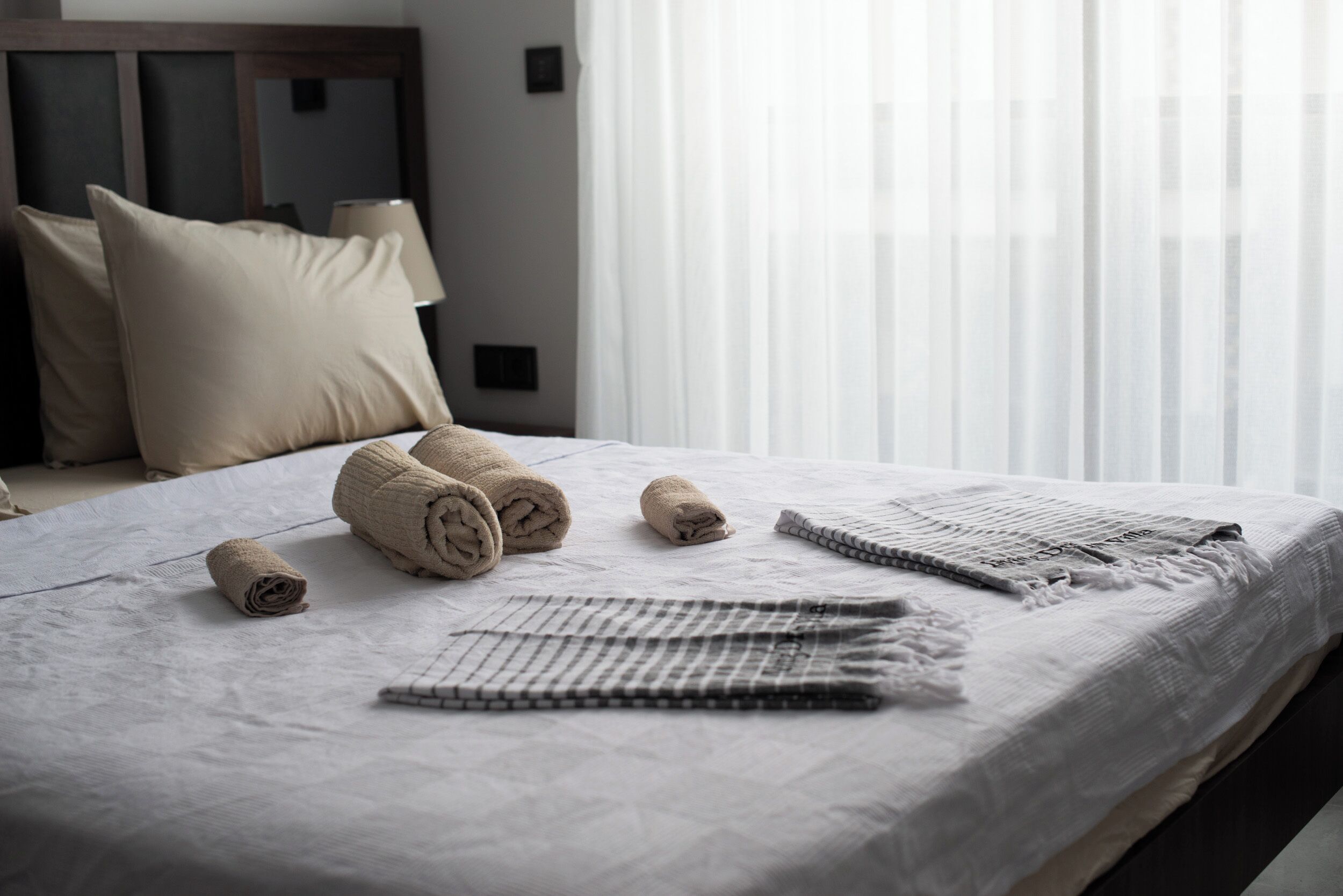 A neatly made bed with rolled towels and a checkered throw, a lamp, and sheer curtains.