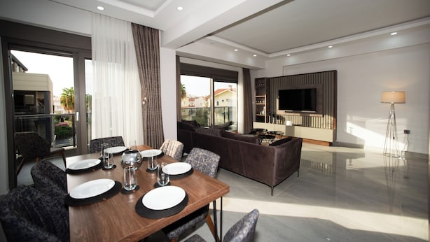 A modern dining area with a wooden table set for four, a sofa, and a TV in the background. - Living Dream Duplex Belek (Antalya)
